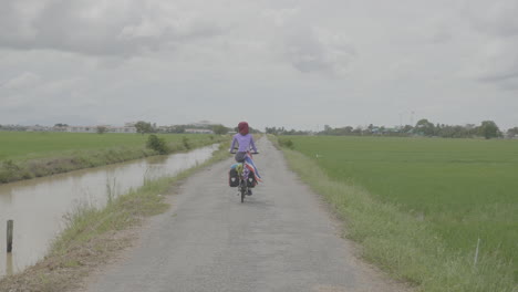 Vista-Trasera-De-Una-Mujer-En-Bicicleta-A-Través-De-Campos-De-Hierba-Abiertos-En-Malasia-Con-Bandera-Tailandesa