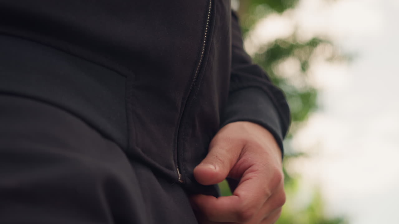 백인 남성이 야외 산책을 준비하고 있어요. 그의 뒤로는 나무들이 보이고, 드로스트링을 고정하는 손이 클로즈업되어 있으며, 야외 활동을 위해 옷을 정리하는 손에 초점이 맞춰져 있어요.