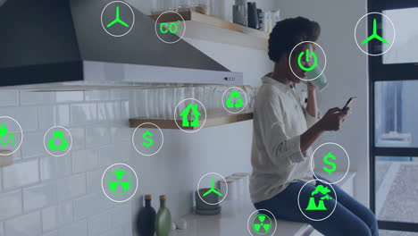 animation of eco icons over african american woman using smartphone in kitchen