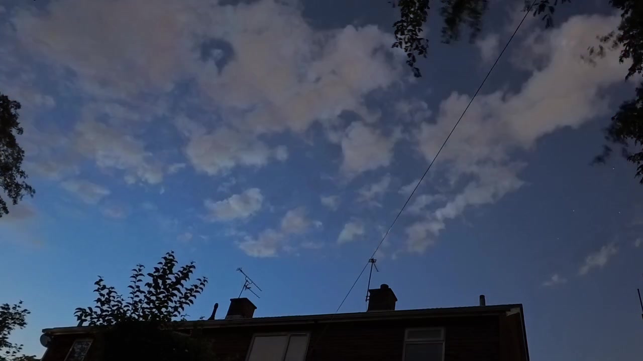 Time-lapse da chuva de meteoros Perseidas sobre o telhado de uma casa residencial, com nuvens de céu noturno em rápido movimento.