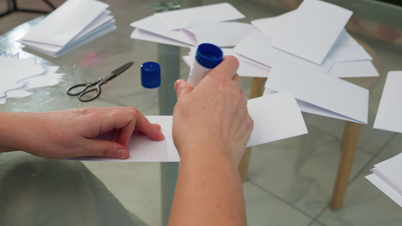 Close-up hand view of applying glue to crafted and folded paper, preparing for next step in paper craft project, with scissors and more folded papers in background