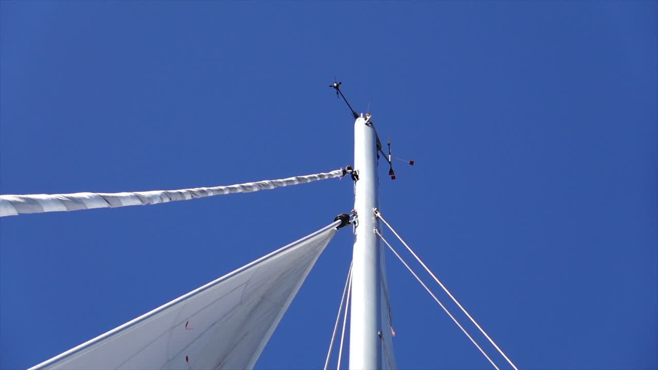 Camera pans to the Mast of a sail boat and head of the sail on a blue sky day.