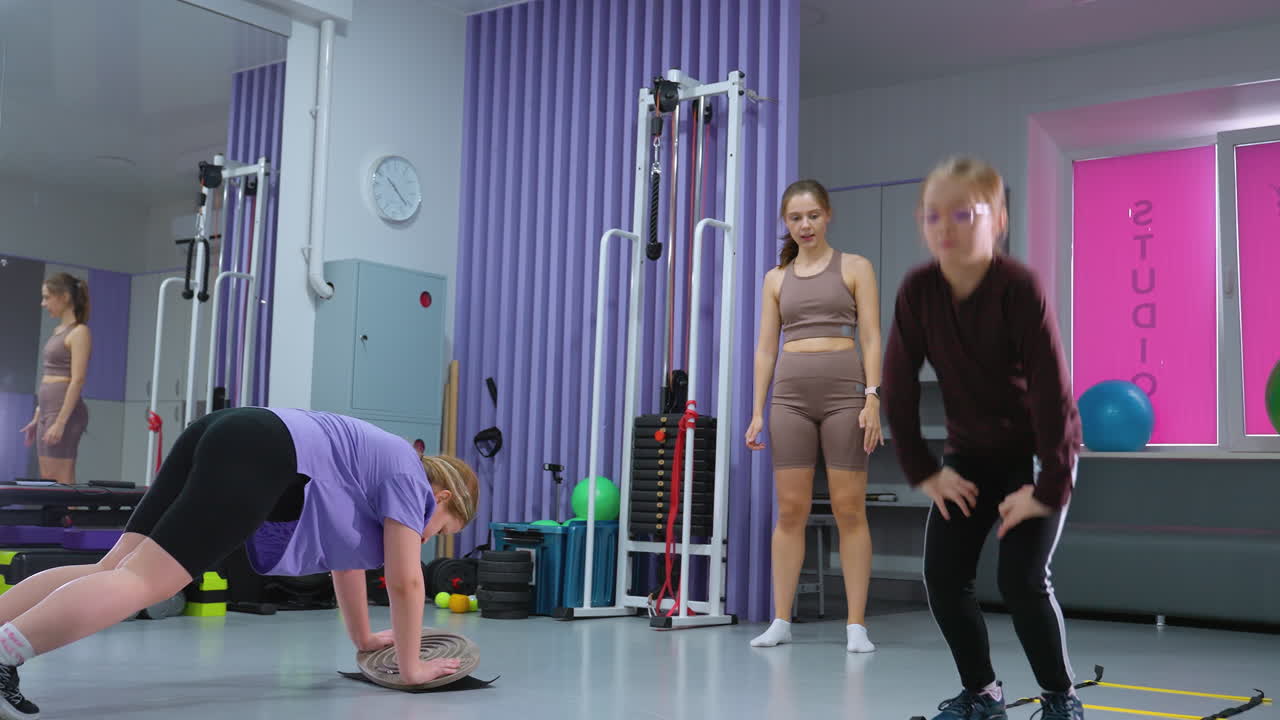 niños involucrados en una sesión de entrenamiento con el profesor monitoreando el progreso, uno equilibrando a bordo, otro saltando una escalera de agilidad, un gran espejo refleja el entorno del gimnasio, equipos de fitness