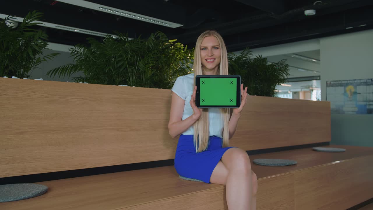 Woman showing tablet in office. Young smiling blond woman showing tablet with green chromakey screen looking at camera and smiling in office