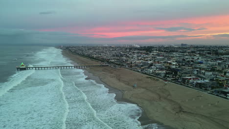 manhattan beach ocean waves with pier overlooking an urban cityscape with morning pink sunrise, aerial drone panning shot