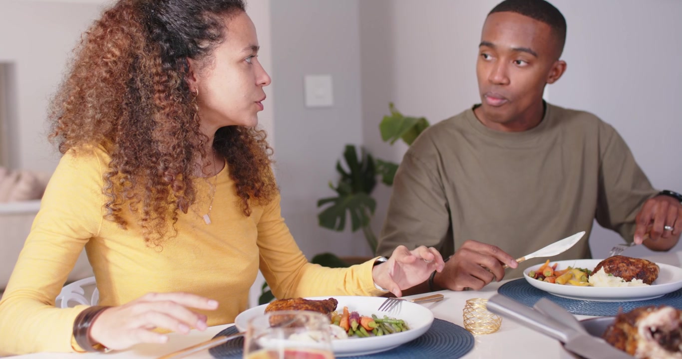 Couple sitting at table cutting food with forks, woman touching man getting attention, man eating