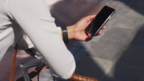 midsection of african american woman using smartphone leaning on bike in street