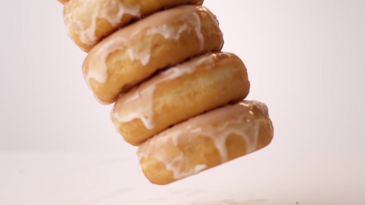 Ciambelle lucide glassate allo zucchero impilate che cadono su un tavolo bianco e danzano in slow motion estremo. Contenuti per la giornata nazionale del donut, pubblicità per vetrine di panetterie, spot pubblicitari per la voglia di zucchero