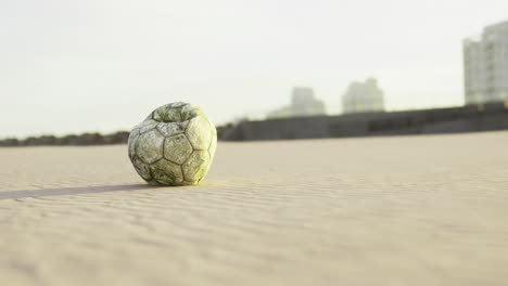 soccer ball rests on sandy beach with city buildings in background