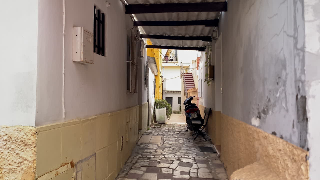 Stretto vicolo in una città spagnola con muri antichi, uno scooter parcheggiato e un tradizionale percorso in acciottolato, che evoca la vita urbana storica.