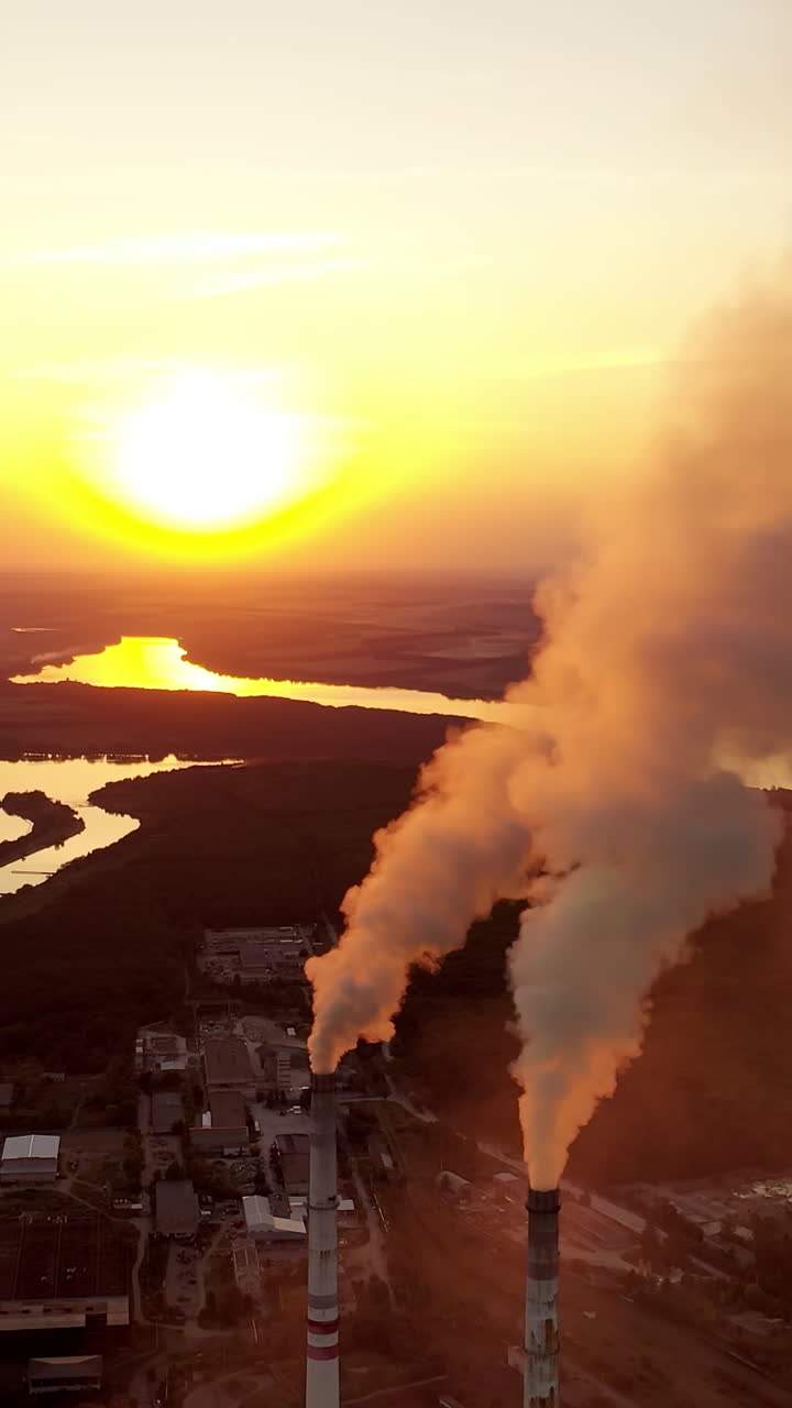 Pipes with smoke near the evening river. Pollution of the environment. Industrial plant at sunset. Dark smoke coming from plant pipes. Vertical video