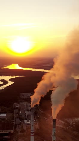 pipes with smoke near the evening river. pollution of the environment. industrial plant at sunset. dark smoke coming from plant pipes. vertical video
