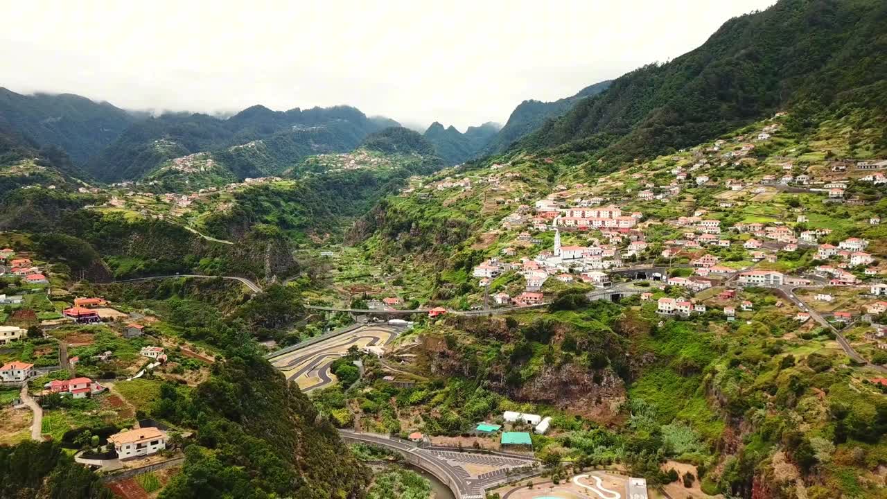 Lush vegetation covers the valley, surrounding the charming village nestled amidst the dramatic slopes, creating a tranquil scene under a cloudy sky in Faial, Madeira, Portugal drone pulling out shot