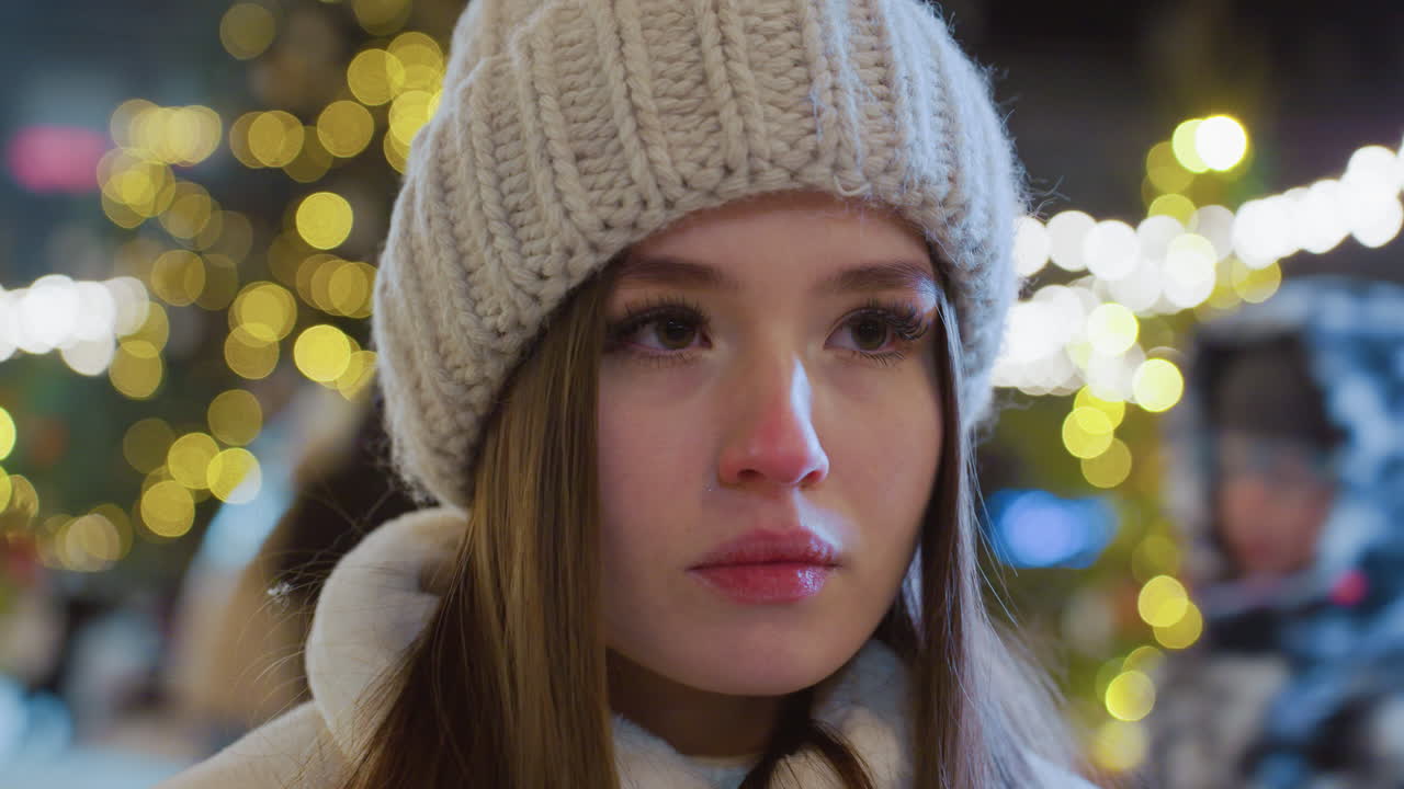 Close-up of young woman wearing a knitted beanie and winter coat, sitting alone with eyes closed in deep reflection, blurred festive city lights and people moving in the background