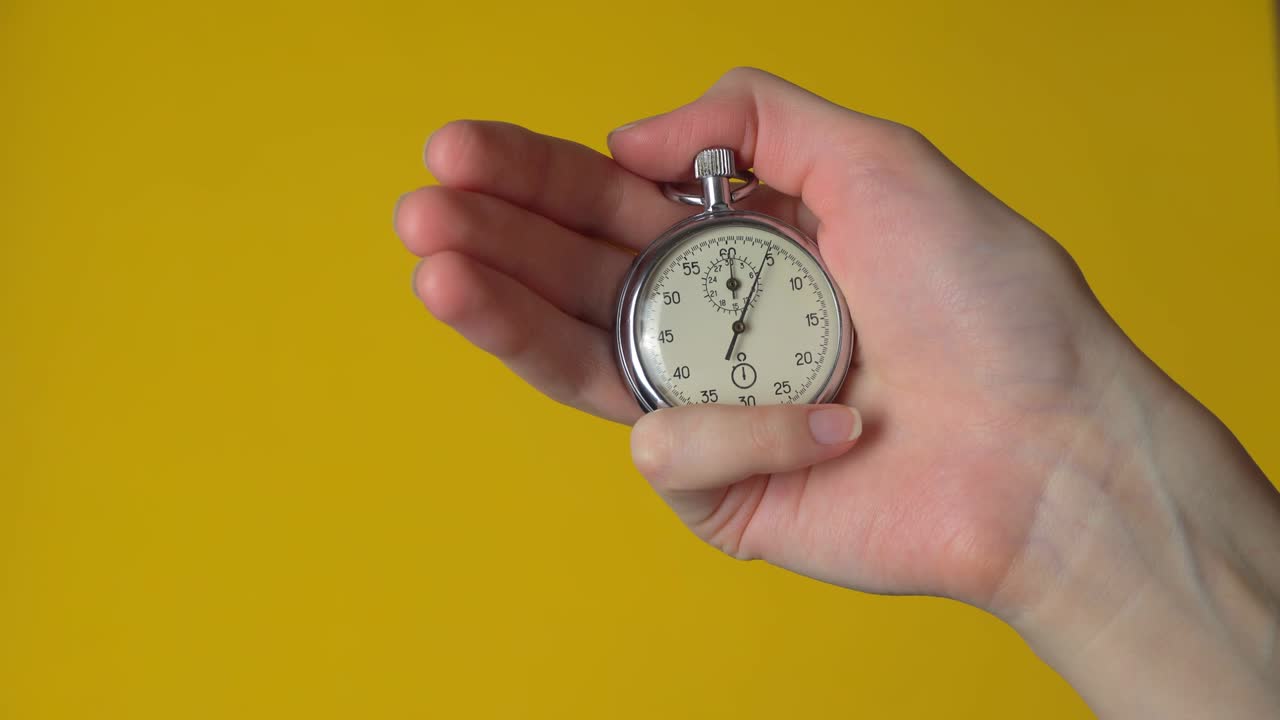 A woman's hand holds an analog stopwatch on a yellow background.