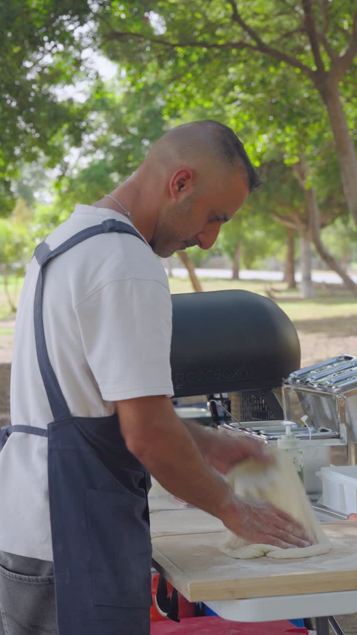 Slapping and turning Neapolitan dough in wooded park setting, cooking moment with Mediterranean pizzaiolo and mobile oven