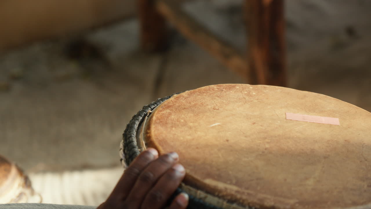 Close-up of African male hands playing a bongo, highlighting the repaired drum, set against the backdrop of the late afternoon's beautiful lighting