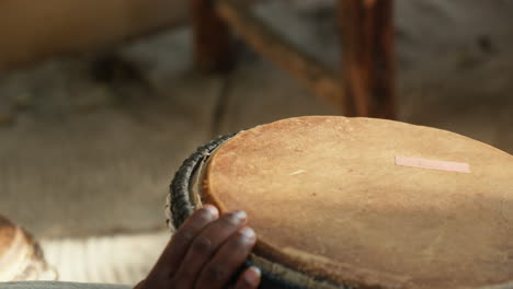 Close-up of African male hands playing a bongo, highlighting the repaired drum, set against the backdrop of the late afternoon&#039;s beautiful lighting