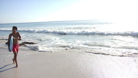 A-young-biracial-man-walks-along-the-shore-with-a-surfboard,-with-copy-space