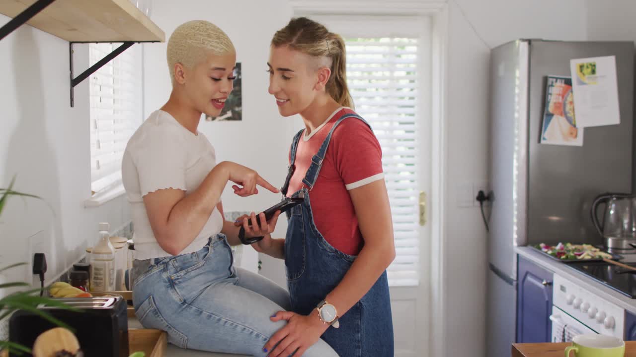 Happy diverse female couple talking and using smartphone together in kitchen
