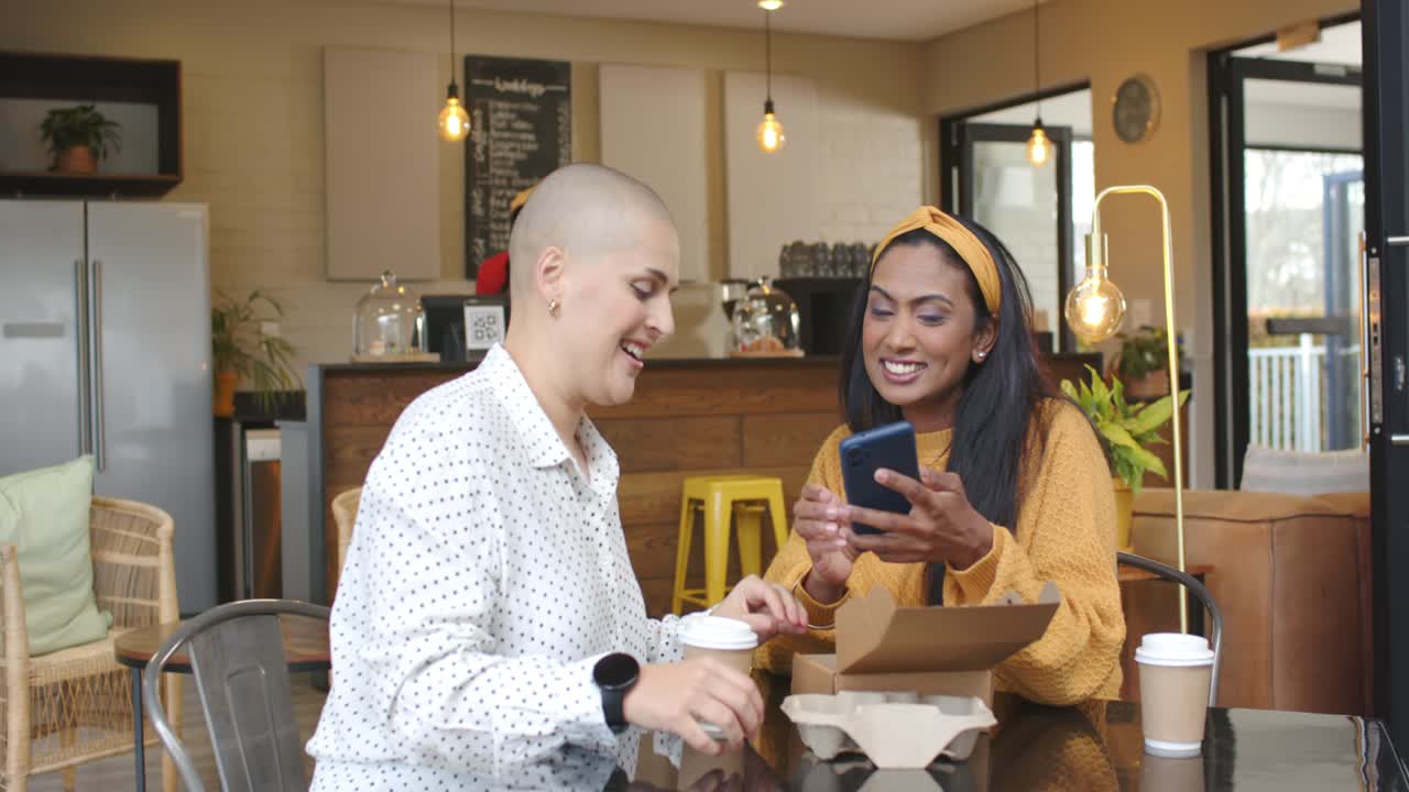 Diverse female friends responding to barista wave, grabbing cup and sharing phone meme at cafe