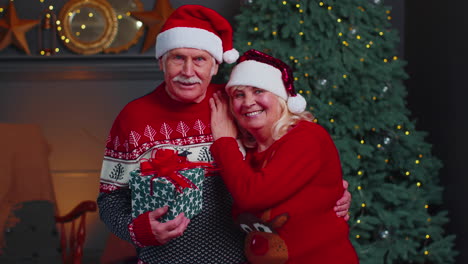 Senior-couple-family-in-Santa-Claus-hats-celebrating-Christmas-looking-at-camera-and-hugging-at-home