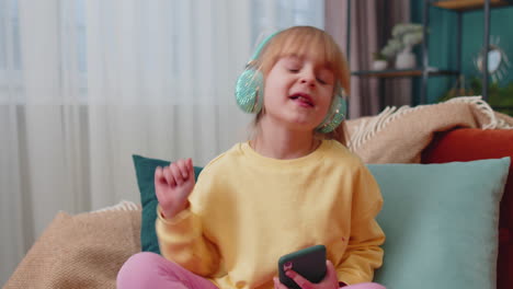 child girl kid with smartphone in headphones dancing singing listening music at home alone on sofa