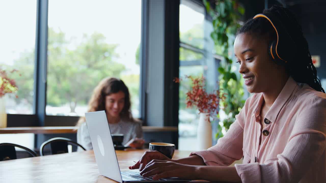 Businesswoman Wearing Wireless Headphones Works On Laptop In Cafe Shop With Colleague In Background