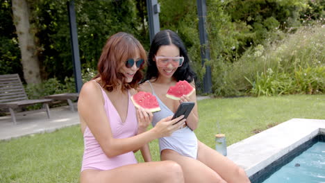 two female friends in swimsuits enjoy watermelon by a pool, one showing something on a phone