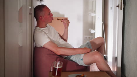 freelancer seated comfortably in hallway savoring turkey meat, relaxing on soft beanbag chair with plate of food and glass of juice on cardboard box beside him