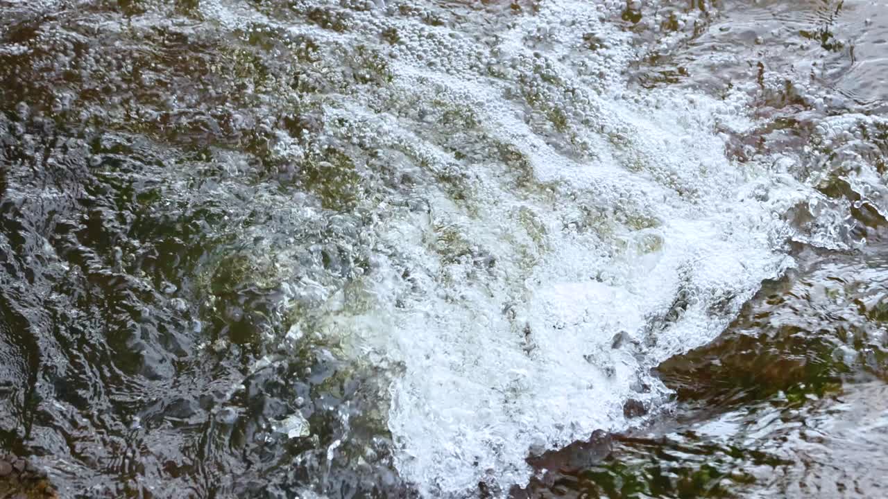 A dynamic stream flows over mossy rocks, creating bubbles and splashes in natural light