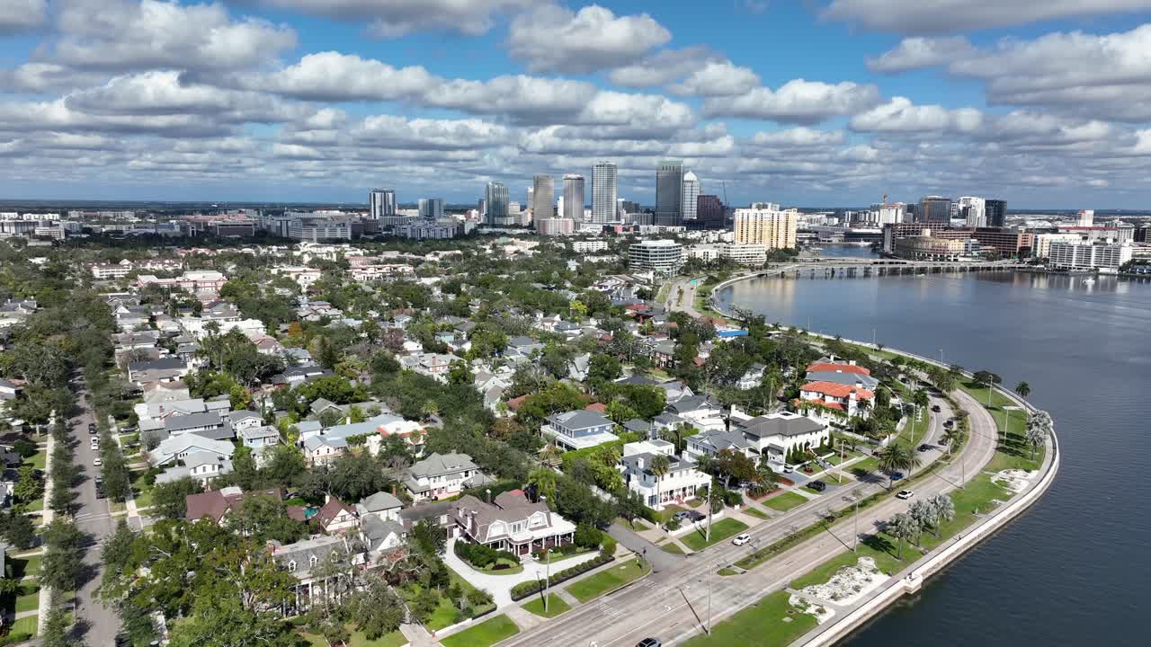 Vista aérea de Tampa, Flórida, mostrando um bairro residencial costeiro com palmeiras, estradas à beira-mar e o horizonte do centro da cidade sob um céu azul brilhante. Grande plano em ascensão. Tráfego na rua ao longo da costa.