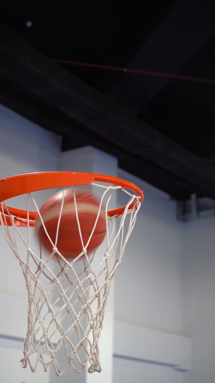 Primo piano cinematografico di un pallone da basket che entra in un canestro arancione con retina bianca durante una partita o una sessione di allenamento al coperto. B-roll sportivo ad alta energia.