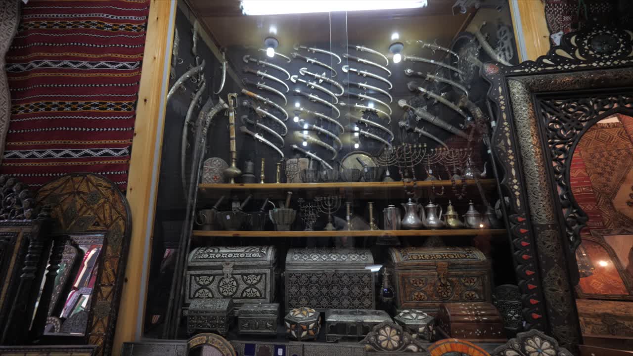 View from left to right of the wall of an antique shop decorated showcasing with variety of silver knives, wooden frame mirrors and filligree pattern trunk boxes.