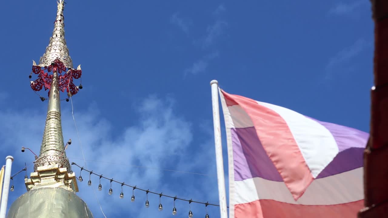 A vibrant Thai flag flutters next to an ornate golden pagoda spire under a clear blue sky.