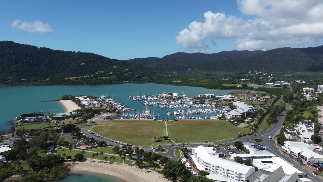 Prospettiva aerea sulla Coral Sea Marina ad Airlie Beach che mostra yacht ormeggiati, parchi costieri e le colline verdi circostanti sotto un cielo azzurro brillante