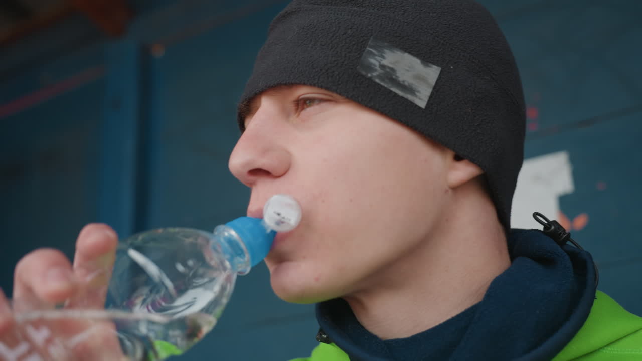 초록색 후드티를 입은 백인 남성이 플라스틱 병에서 물을 마시고 있어요. 캡과 턱선이 보이는 강렬한 클로즈업 샷으로, 골대 근처에서 추운 날씨에 야외 훈련 중 휴식을 취하며 캐주얼 비니를 쓰고 스포티한 분위기를 연출하며 빠르게 수분을 보충하고 있습니다.