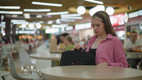 lady wearing black crop top under pink shirt sits in restaurant, opens black bag, and removes laptop and placed on the table, the well-lit restaurant has blurred background of people dining