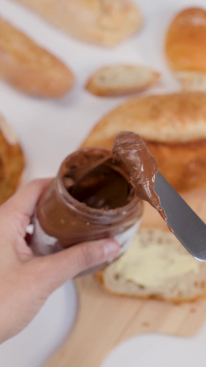 Mano che preleva crema spalmabile al cioccolato e nocciole da un barattolo con un coltello.