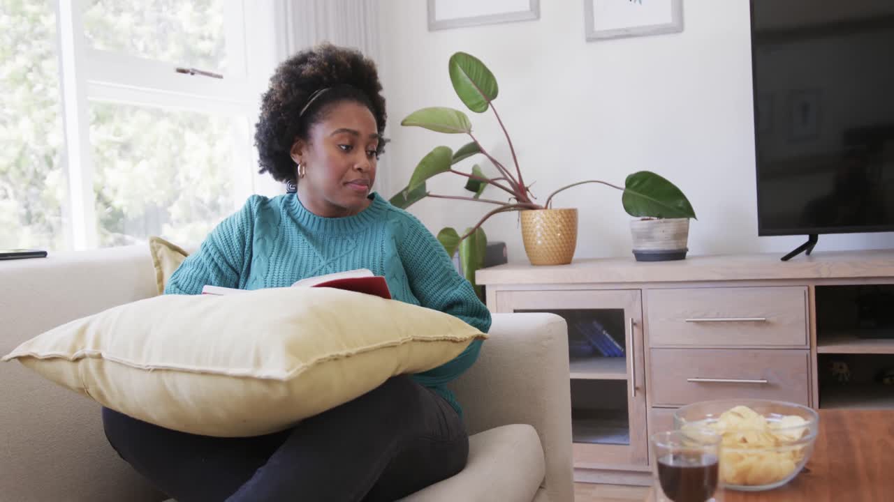 Happy african american woman relaxing on couch reading book and eating chips, slow motion