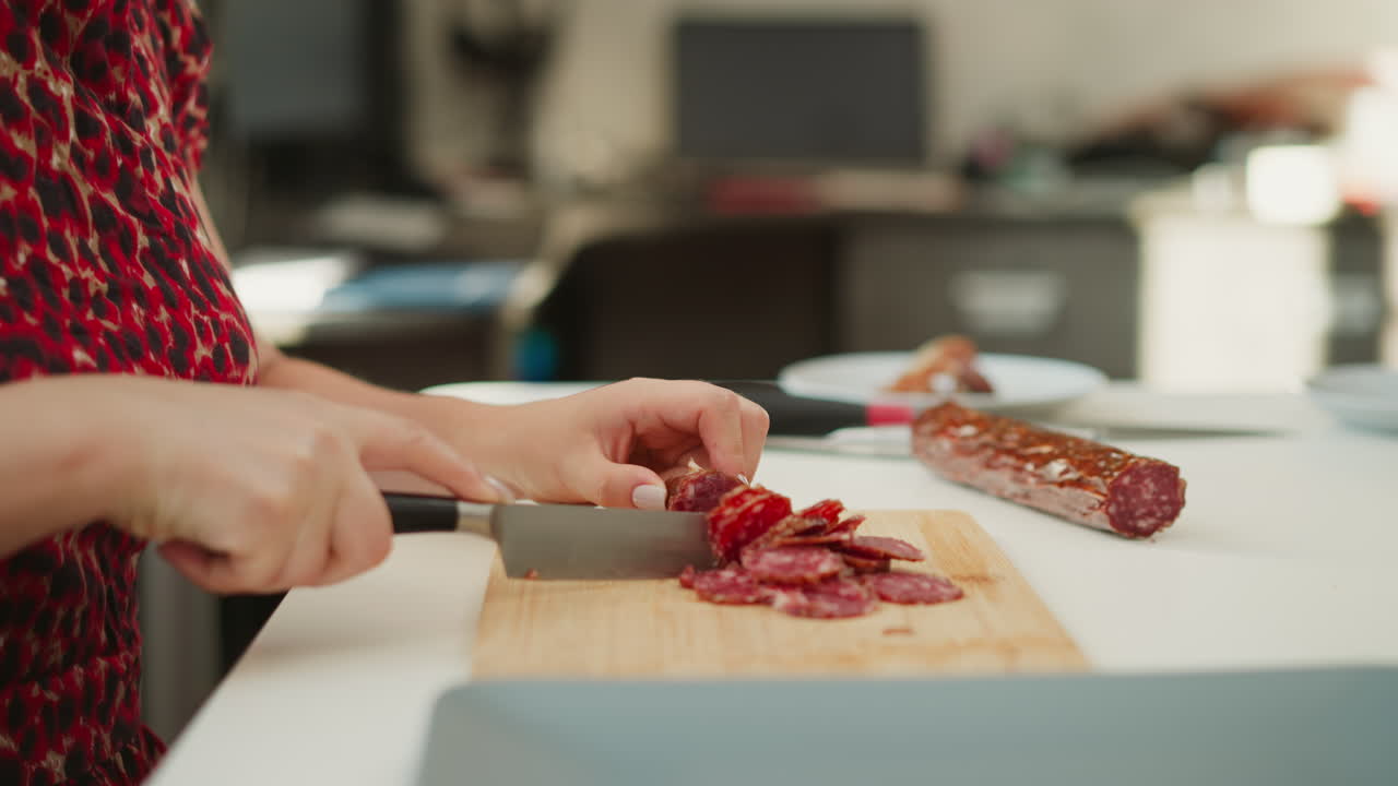 Cook hands slicing cured pork sausage into thin round pieces with sharp chef knife on wooden board over white table by bright window, highlighting marbled texture and precise culinary technique