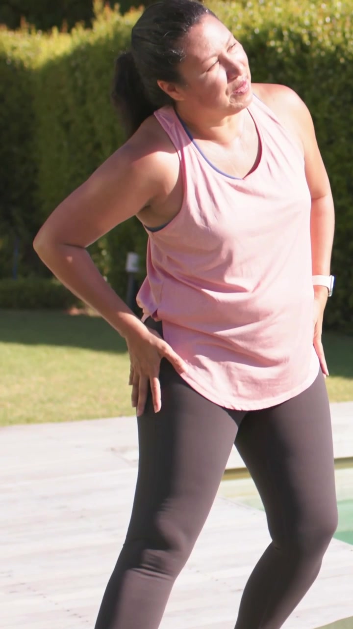 Vertical video: Leaning, sliding hand down right thigh woman in pink tank warming up by pool on mat