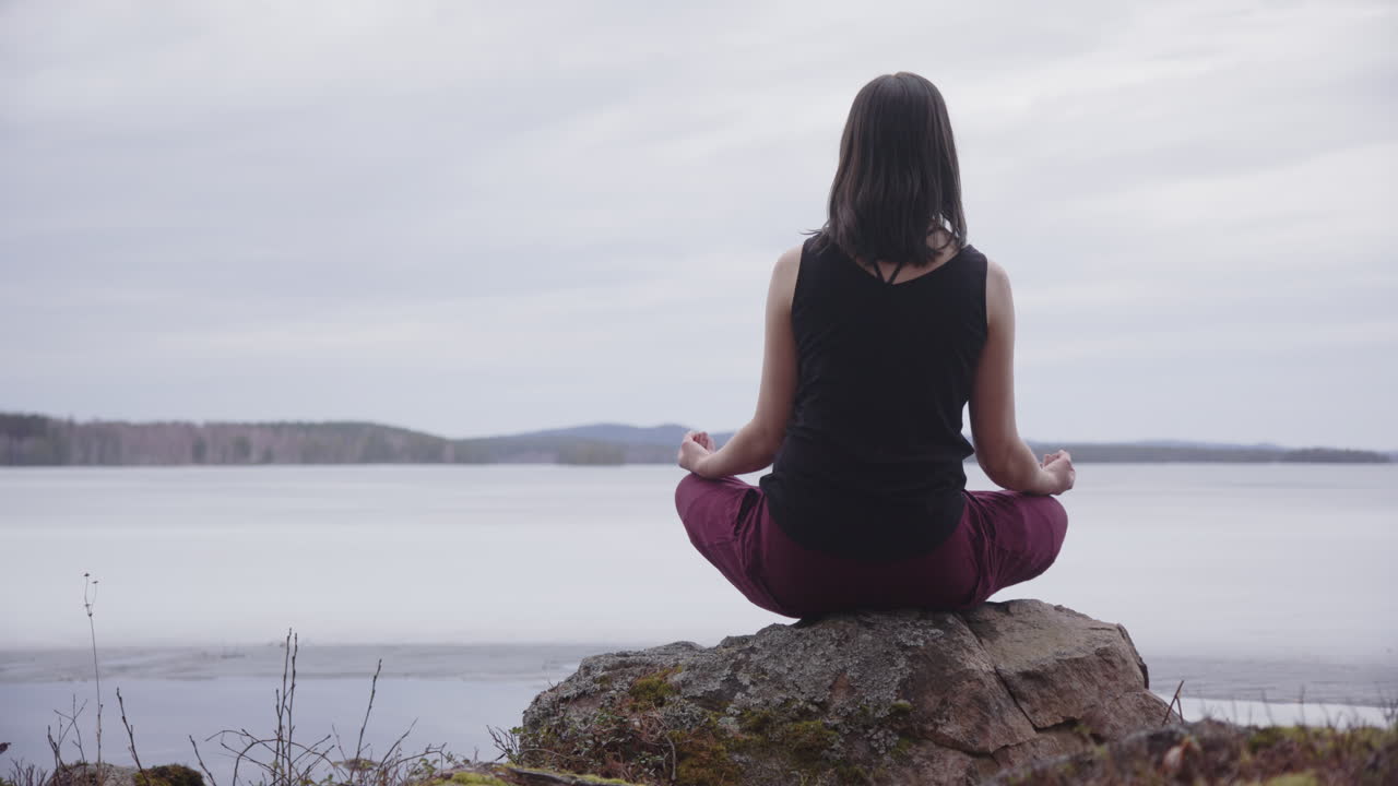 una dama sentada en posición de loto en una roca medita con vistas al lago congelado, sweden