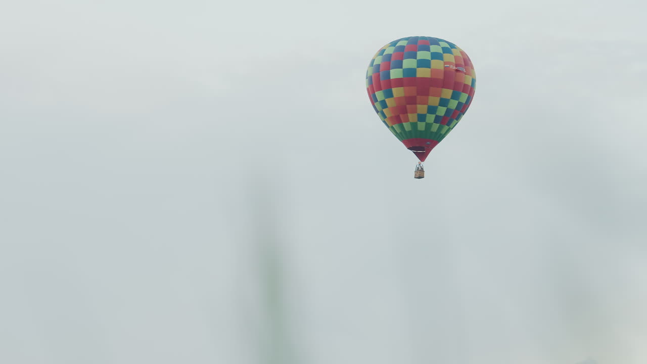 blurred foreground grass framing colorful checkered hot air balloon with passengers rising high above misty countryside sky during peaceful dawn journey showcasing solitary distant balloon