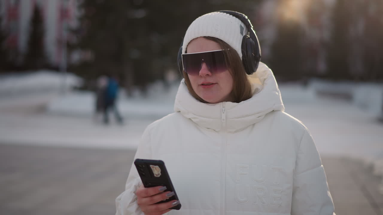Performer pressing mobile phone and swaying to beat in busy snowy park at sunset with passersby in background, wearing sun glasses, headset, white coat and beanie glowing in warm backlight