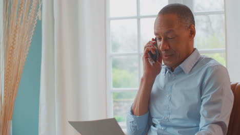 mature man or businessman with mobile phone working from home in armchair looking through paperwork