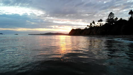 drone aéreo disparado al atardecer volando bajo sobre el agua y reflejos de la superficie del océano pacífico con la silueta de los acantilados de la costa de california y palmeras en santa barbara, california