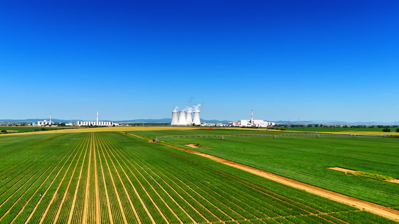 Fields under a clear blue sky. Green fields stretch across the landscape with industrial plants in the background beneath a sunny sky