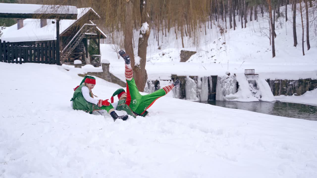 Two funny elves in green costumes playing happily on winter background. One dwarf falling into white snow near the river. Happy Christmas holidays.