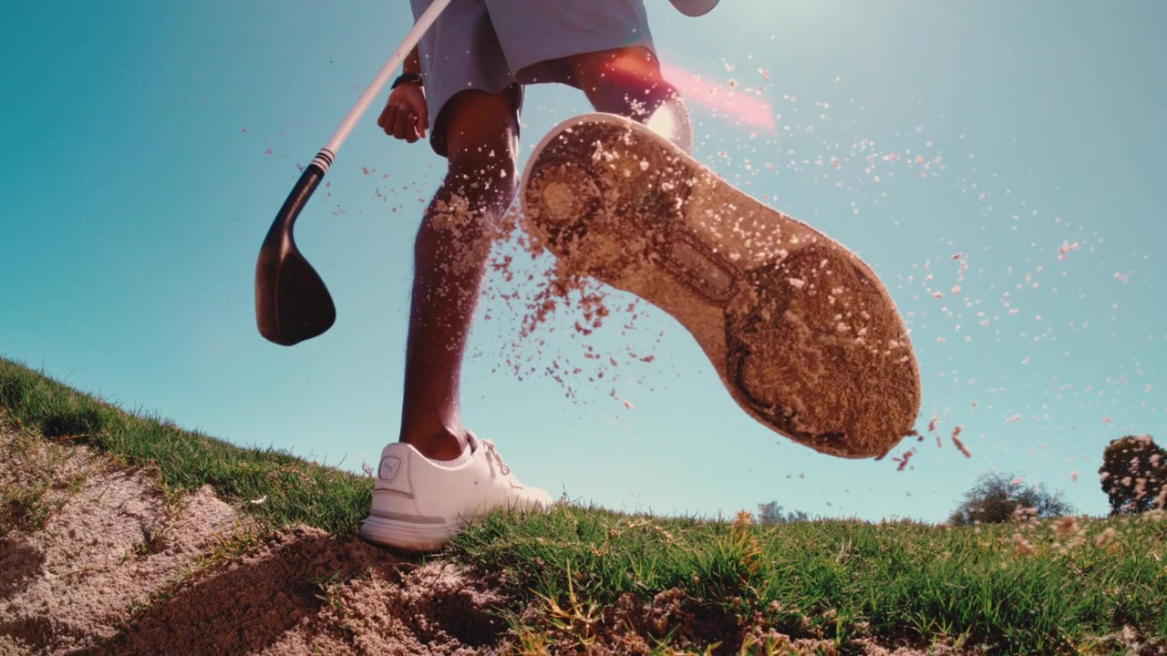 A low angle follows a golfer's legs and white shoes walking through a sand bunker, cleaning sand off cleats with wedge impact off shoes in 4k 800fps slow motion on sunny day